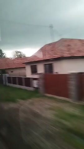 The video shows a blurry, fast-moving view of houses with red tile roofs from the perspective of someone inside a vehicle. The houses appear to be in a rural or suburban setting with fences and greenery. The sky is overcast. There are brief glimpses of a car and what appears to be a picture on a window. The motion blur suggests the vehicle is traveling at a moderate to fast speed.