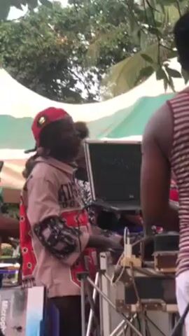 The video shows a DJ working on a laptop, with speakers and other equipment nearby. He is wearing a red cap, a patterned shirt and has dreadlocks. People are standing around him, some recording with their phones, dancing, and chatting. A large tent is set up in the background, and there are trees overhead. The scene suggests an outdoor event or party.