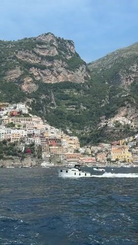 The video shows a beautiful view of Positano, Italy from the sea. A white motorboat speeds across the water.  Positano's colorful houses cascade down the hillside towards the sea. The backdrop is a rugged mountain covered with greenery. The sky is blue with some clouds.