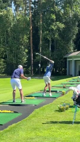 This video shows a golf driving range on a sunny day. Two men are visible on separate practice mats, both in the process of swinging golf clubs. The man in the foreground on the left is wearing a blue polo shirt and white shorts. The man on the right is wearing a blue long-sleeved shirt and white shorts. Several rows of golf balls are visible on the mats, and baskets of golf balls are placed on the grass. Golf bags are also visible on the right side of the frame. In the background, a building with large windows and the words "Russian Golf Academy" are visible. Other golfers and instructors can be seen further down the driving range. The environment is a grassy area surrounded by trees.