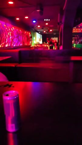 The video, shot from a low angle, shows the interior of a nightclub. In the foreground, a dark table surface is partially visible, with a can of a beverage resting on it. The lighting is dim and vibrant, alternating between red, blue, and purple, indicative of a nightclub atmosphere. The background features tiered seating booths with red lighting underneath. In the distance, two individuals are seen near a brightly lit bar area, seemingly dancing or gesturing. The room has decorative elements, including a wall feature with what appears to be a pattern of abstract shapes illuminated with various colors. The music plays loud in the background, and the camera is unsteady.