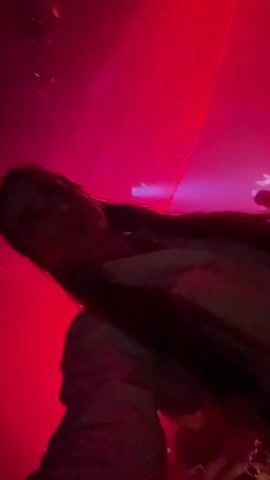 A woman with long, dark hair stands in a crowded nightclub, filming a selfie video. She's wearing a white shirt. The club is filled with fog and colorful lights, mostly pink and green. Other people can be seen dancing in the background. She smiles and looks directly at the camera. The camera angle is tilted upwards, providing a low-angle view. The video appears to be taken in a dimly lit, energetic environment.