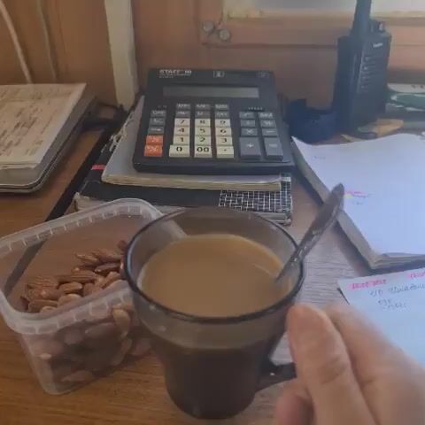 The video opens with a close-up of a mug of coffee with a spoon in it. A hand is visible holding the mug. The camera then pulls back to reveal a desk setting. On the desk, there is a plastic container filled with almonds, a calculator, a stack of papers, and a laptop. The laptop screen displays the words "Электронное сольфеджио," which translates to "Electronic Solfeggio." The background appears to be an office or study environment with wooden walls. The lighting suggests it is daytime.