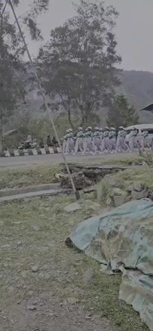 A group of people dressed in matching green and white uniforms are marching in formation along a paved road. They are walking in rows and appear to be participating in a parade or drill. A flag, white and red, is visible on a pole to the left of the marching group. The environment is outdoors with trees and hills in the background. The road is bordered by grassy areas and some rocky terrain. There is a camouflage-patterned tarp or fabric in the foreground. A white van is parked on the right side of the road, behind the marching group. There are a few other people standing on the side of the road watching the procession.