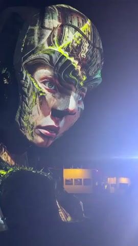 This video shows a projection mapping art installation at night. The projection features a large, detailed face with green and brown abstract patterns superimposed onto it. In the background, there are some lights and what appears to be a display of framed images. The projection is cast onto a large sculpture of a hand that is illuminated in purple light. The overall scene suggests an outdoor art exhibition or event.