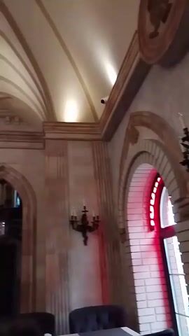 The video shows the interior of a building with ornate architecture, including arched doorways, decorative molding, and patterned ceilings. The building has multiple levels, with the upper level featuring a balcony overlooking the lower area. A chandelier with red and white elements hangs from the ceiling. The lighting is a mix of natural and artificial light. There is furniture present, including what appears to be chairs. People are visible on the upper level.