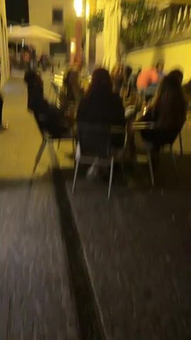 The video captures a group of people seated at an outdoor cafe, while another woman walks by on a cobblestone street. The cafe appears to be located in a narrow alleyway, illuminated by warm streetlights. The seated group consists of several individuals, some engaged in conversation with drinks on the table. A woman in a red sweater and light blue jeans walks from right to left, passing the cafe. The background reveals more outdoor seating areas, with additional patrons under umbrellas. An "ATM" sign is visible on a building in the distance. The overall atmosphere suggests a relaxed, evening scene.