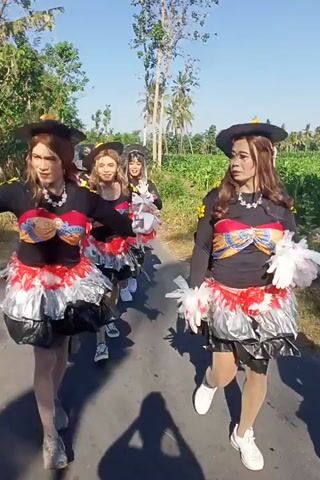 The video shows a group of people dressed in colorful costumes, possibly for a parade or performance. They are outdoors, with trees and a road visible in the background. The people are wearing elaborate outfits with sequins, feathers, and large bows. Some are wearing hats, and all have makeup on. The camera appears to be handheld, and the footage is shaky at times. The people are smiling and interacting with each other, suggesting a festive atmosphere.