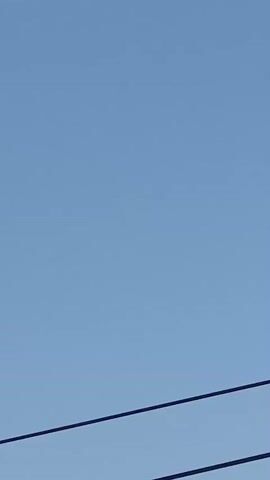 The video shows a clear blue sky with two dark, diagonal lines that appear to be power lines or wires in the foreground. In the middle distance, a small, white airplane is visible flying horizontally across the frame from left to right. The airplane is the only discernible object in the sky apart from the wires. The lighting suggests it is daytime.