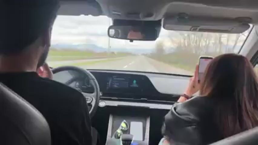 Here is a description of the video:
The video is taken from the backseat of a car driving on a straight, paved road. The driver is a man with dark hair, wearing a black shirt, and is focused on the road ahead. The front passenger is a woman with long, dark hair, wearing a black leather jacket. She is holding her smartphone, presumably taking a video or picture of something outside the car. The dashboard is visible with a digital display showing music information. There are sunglasses and a phone in the center console. Outside the car, there is greenery on either side of the road, and mountains in the distance. A road sign is also visible. The sky is overcast. The people in the car appear to be having a conversation.