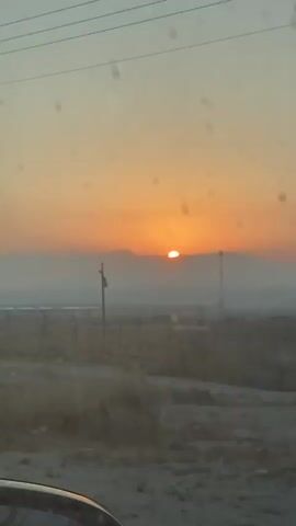 The video shows a sunset over a dry, open landscape. The sun is low on the horizon, partially obscured by clouds or mountains. There is a fence in the foreground, and some utility poles are visible in the distance. The sky is a gradient of orange and yellow. The camera appears to be inside a vehicle, as evidenced by the reflection in the lower left corner of the frame. The overall impression is one of a rural or undeveloped area.