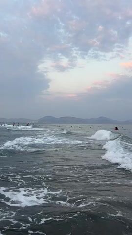 The video shows a body of water with waves and foam. In the distance, there are mountains and a hazy sky with clouds. Several people are visible in the water, some appearing to be swimming or engaging in water activities. The overall scene depicts a coastal environment, possibly a beach, during a time of day when the light is subdued, suggesting it could be dawn or dusk. The focus is on the movement of the water and the distant landscape.
