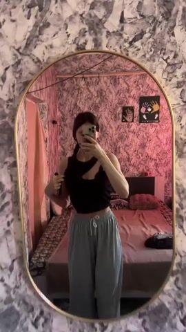 A woman is standing in front of a mirror, taking a selfie. She is wearing a black crop top and grey pants. She has her hair in a braid and is holding a phone in her right hand. The room is decorated with pink wallpaper and has a bed in the background.