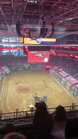 The video captures an indoor monster truck event. A monster truck has overturned on the dirt arena floor. A person in an orange uniform is attending to the monster truck, appearing to attach a winch or strap. Another person in a yellow safety vest stands a short distance away, observing. A yellow construction vehicle, possibly a loader, is positioned near the overturned truck, seemingly assisting. The arena is filled with spectators, and large screens display event information, including a live feed of the monster truck action. The overall environment is that of a large, enclosed stadium set up for a sporting event.