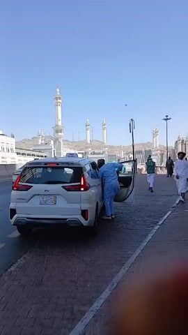 The video shows a white taxi parked on a brick-paved street in front of what appears to be a mosque or other religious building complex.  A man in light blue clothing is standing by the open passenger door of the taxi. In the background, there are several minarets and a large building, possibly part of the religious complex. People are walking on the sidewalk nearby, many dressed in traditional Muslim attire. The sky is clear and blue. The camera is positioned at street level. Some parts of the video are obscured by what appears to be a finger in the bottom right corner.