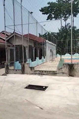 The video begins with a shot of a house under construction. A cement mixer is visible on the left side of the frame. The camera then pans to show a yard with some trees and a volleyball court in the background. A woven basket is placed on the ground near the house. Further along, a yellow truck is seen driving down a dirt road. The video then shifts to show a lush green area with a small house and trees. The camera continues to pan, revealing more of the house and the surrounding vegetation. The video ends with a final shot of the house and its surroundings.