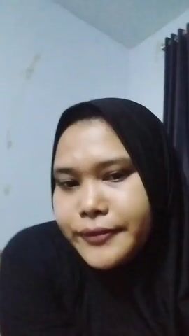 A close-up, slightly blurry video shows a person wearing a black hijab and black clothing. The person's face is partially obscured by their hand, which is brought to their mouth and chin. The background consists of a plain, light-colored wall with some marks, and a dark curtain on the right side. The lighting is dim.