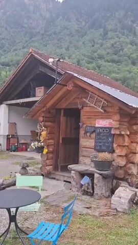 A vertical video pans across a rustic log cabin nestled against a backdrop of lush green mountains and a cloudy sky. The cabin features a wooden door, windows, and a metal roof. A sign on the cabin reads "VENDO" (Italian for "for sale"). To the right of the cabin, a paved path winds through a grassy area with a dark-colored SUV parked on the side. The camera continues to pan, revealing more of the mountainous terrain and a white van parked inside a garage-like structure. The video ends with a view of the interior of the garage, showing the white van and a red piece of machinery.