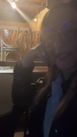 The video shows an indoor scene with people socializing. The first shot is of a woman smiling at the camera. She's wearing glasses and a black jacket over a light-colored shirt. Behind her is a storage area with chairs stacked and equipment lined up. The camera then moves to show a person sitting at a small round table with a glass of beer or wine and a vape device. The scene shifts, capturing multiple people seated at tables, talking. In the foreground, there's a close-up of a glass of beer or wine. The lighting in the video is dim, and the atmosphere appears relaxed.