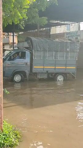 A gray truck is driving through a flooded street. The water level is up to the truck's wheels. The truck has a canvas cover over its cargo bed. There are trees and buildings in the background. The sky is not visible.