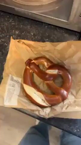 A person is shown adding salt to a pretzel on a paper bag. The pretzel is brown, with a lighter colored portion visible along one side.  A clear plastic container with a white lid is held over the pretzel, and shaken to disperse the salt.  The background includes a black speckled countertop and a stainless-steel appliance, possibly a microwave. A person's legs in blue jeans can be seen in the lower part of the frame.  The hand holding the salt shaker has tattoos on it.