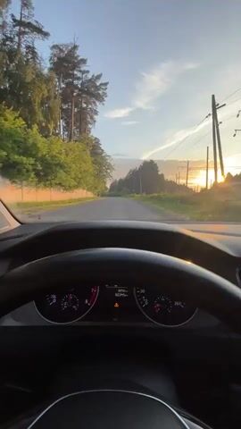 This is a first-person perspective video from inside a car. The driver's hands are not visible, but the steering wheel is in the foreground. The dashboard shows the speedometer, tachometer, and odometer. The car is moving on a road surrounded by trees and vegetation. Power lines are visible on the right side of the road. The sun is setting, casting a warm glow and long shadows. The sky is blue with some clouds. The road appears to be paved and leads into the distance. There are a few other cars visible on the road.