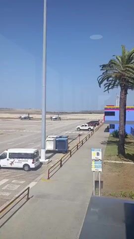The video shows an airport tarmac scene on a bright, clear day. Two small planes with white and blue markings are visible, stationary on the tarmac. A white van labeled "FBO Caribbean" and a dark-colored passenger van are parked nearby. An airport stairway marked "AEROPOSTAL" is also present. The backdrop is a flat landscape meeting the horizon under a cloudless blue sky. The video appears to be shot from inside a building, looking out through a window. The scene feels calm and static, with no immediate activity taking place.