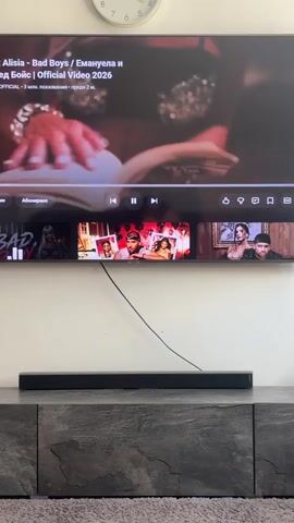 The video shows a television screen displaying a music video. The video is paused, and a progress bar with thumbnails is visible at the bottom of the screen. The thumbnails show different scenes from the music video, including shots of women in various outfits and poses. The television is mounted on a wall above a media console. A soundbar is placed on the media console. The room appears to be dimly lit.