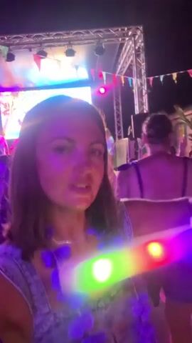 A woman with medium-length brown hair is recording a selfie video at night outdoors at what appears to be a festival or concert. She is wearing a denim outfit and a purple flower lei. She moves her head and mouth as if speaking or singing. In the background, there is a stage with bright lights and colorful flags strung across it. Other people are visible in the background, some also appearing to be at the event. There are also some blurry lights in the foreground, possibly from other attendees or decorations.