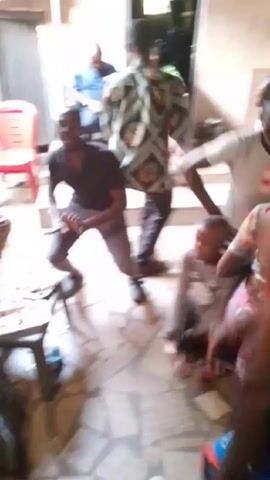 The video shows a group of people, including a young child, dancing indoors. Several adults are moving and grooving to music, while the child stands mostly still, occasionally being touched or guided by an adult. The setting appears to be a casual indoor space, possibly a home, with basic furnishings. The room is somewhat dimly lit, and the focus is on the lively interactions and dancing. The video perspective is handheld, moving with the action, which causes some blurriness.