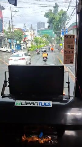 The video appears to be filmed from inside a vehicle, likely a bus or a jeepney, looking out the windshield at a street scene. The windshield has two windshield wipers, one of which is in the lowered position and the other is partially raised. There is a black screen or monitor mounted on the dashboard in front of the viewer. A green and white "cleanfuel" sticker is visible on the dashboard. Outside, it is daytime and appears to be raining or has recently rained, as the road is wet and reflects the surroundings. There are several vehicles on the road, including a white car, several motorcycles with riders, and a jeepney. The jeepney has text on its side that reads "GABBY S KALIWA" and some other illegible text. Buildings and trees line the street, and utility poles and wires are visible. In the distance, there are more buildings, some of which appear to be under construction. The sky is overcast. A blue electric fan is mounted on the dashboard, pointing upwards.