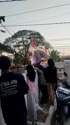 This video shows several people, including children, gathered around a large effigy. The effigy appears to be a monster or demon-like figure with a large head, white hair, and red horns. Some people are touching or interacting with the effigy. In the background, there is a tree, buildings, and a motorcycle parked on the side of the road. The sky is overcast, suggesting it might be dawn or dusk. The video is shot from a slightly elevated angle and is oriented vertically.