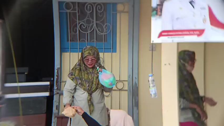 A woman wearing a hijab and glasses stands in front of a doorway. She is holding a colorful plastic bag and a brown paper bag. A young person wearing a hijab kneels in front of her, reaching for the brown paper bag. Another woman in a hijab stands to the right of the first woman. A banner hangs on the wall behind them. A plastic bottle hangs from a string on the wall.