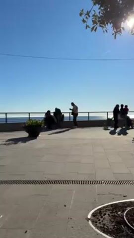 The video shows a plaza or walkway in front of an ocean view, under a clear blue sky. Four people are standing near a railing overlooking the ocean, while another person walks across the plaza. The plaza is paved with square grey stones. A planter sits to the left of the group. The camera remains stationary.
