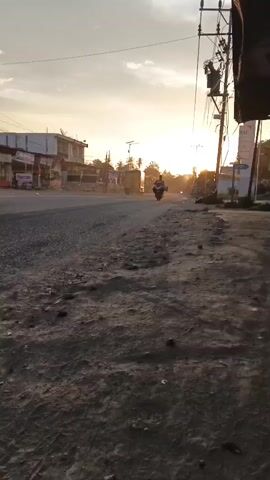 A low-angle, ground-level shot captures a dirt and gravel road adjacent to an asphalt road. In the distance, at the vanishing point, a motorcycle is visible with a rider. The sun is setting, casting a warm glow over the scene and creating a bright haze. Buildings line the left side of the asphalt road, and utility poles and wires are visible on the right. The overall atmosphere suggests a rural or semi-urban setting at dusk.