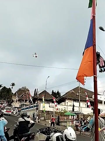 The video shows a street scene with several motorcycles parked in the foreground. In the background, there are buildings, people walking, and a kite flying in the sky. The overall atmosphere appears to be festive, with flags and decorations visible. The sky is overcast.