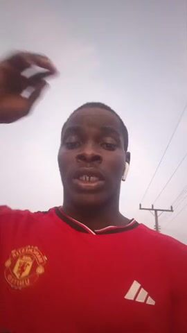 A man is recording a video of himself while walking outside. He is wearing a red Manchester United jersey and white earphones. He touches the back of his head, and looks up at the sky, speaking but the audio is indistinct. The sky is cloudy and there are powerlines visible in the background.
