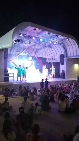 A group of people are sitting on the ground watching a performance on a stage. The stage is lit with colorful lights and has several performers on it. One performer is wearing a green shirt and shorts, and another is wearing a yellow skirt and a white top. There are also some large inflatable spheres on stage. The audience is made up of people of all ages, and they appear to be enjoying the show. The event is taking place at night, outdoors, under a large covered structure.