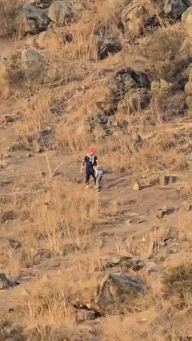 A video shows a wide shot of two people walking down a hill covered in brown grass and rocks. The person in front is wearing a blue and black outfit and a bright red hat. They are holding the hand of a child who is walking next to them. The hill is sparsely vegetated and dotted with rocks. In the background, more of the hill can be seen, with a few trees at the top.