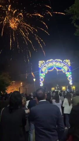 A nighttime outdoor scene features a crowd of people watching fireworks. The fireworks are exploding in various colors, primarily orange and red, against a dark sky. In the foreground, the backs of several people are visible, facing away from the camera. To the right, a brightly lit archway decorated with blue and white lights stands prominently. In the background, trees and some structures are dimly lit. The overall atmosphere suggests a festive event or celebration.