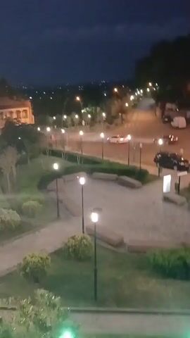 The media file shows an aerial view of a park area at dusk. The scene is dimly lit, with streetlights casting a warm glow on the pathways and surrounding greenery. In the foreground, there are paved walkways, benches, and manicured bushes. Beyond the park, a street is visible with several cars driving by, their headlights and taillights creating streaks of light.

Further in the background, there are buildings with illuminated windows and more streetlights stretching into the distance, suggesting a town or city. The sky is a deep blue, transitioning from daylight to night, with some clouds visible. The overall atmosphere is peaceful and quiet, characteristic of early evening. There is no visible text in the media file.