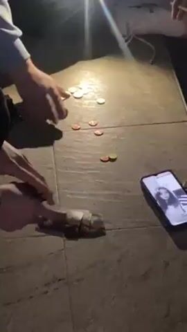 Two people are gambling on the floor, with loose cash and coins on the floor in front of them. The floor is covered with a light-colored, square-tiled surface. A phone is laying nearby with a woman’s face visible on the screen. A light source is shining brightly nearby, casting harsh shadows. One person’s foot is visible, wearing a white shoe and sock.