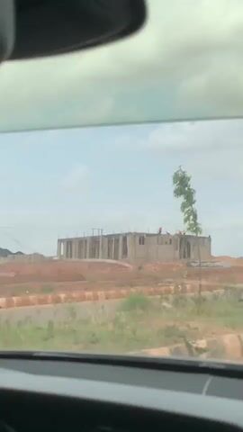 From inside a car, the video shows a view of a construction site in a dry, grassy, rural area. The sky is partly cloudy. A two-story concrete building is under construction. There are a number of workers on the roof, visible by their bright colored shirts. A small tree is in the foreground and blocks the view of part of the building. The car has a crack in the windshield. Upbeat music is playing.