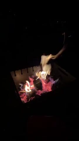 The video shows a close-up of a campfire in a metal fire pit at night. The fire is burning brightly with orange and red flames, illuminating the dark surroundings. There are logs or branches visible within the fire pit, and embers glow red beneath the flames. The fire pit has a perforated metal edge. The background is completely dark, with a few distant, indistinct lights visible in the upper portion of the frame.