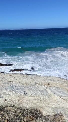 The video shows a rocky coastline with waves crashing against the rocks. The water is a gradient of blue, from a lighter turquoise closer to the shore to a deep navy blue further out. The sky is clear and bright blue. The rocks are light gray in color. There is some vegetation visible in the foreground. The waves are white and foamy as they break against the rocks.