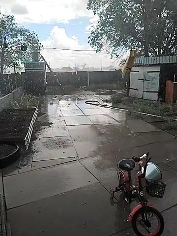 This video captures an outdoor scene during what appears to be a rain shower. A patio area paved with large square tiles is wet, indicating recent or ongoing precipitation. In the foreground, a small pink bicycle with training wheels and a basket is parked. To the left, there is a raised garden bed filled with dark soil, bordered by a wooden fence. In the background, a chain-link fence encloses the property. A simple shed-like structure with a light-colored wall and a darker roof is visible on the right. Trees with green foliage are present in the background, some with branches extending into the frame. The sky is partly cloudy, with patches of blue sky visible amidst the gray clouds, suggesting variable weather conditions. There are no visible people in the video.