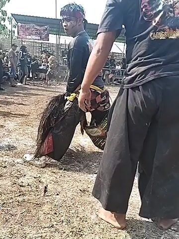 The video shows a traditional Indonesian performance taking place outdoors on a dirt surface littered with leaves. A young man, dressed in black pants, a black long-sleeved shirt, and white shoes, is wearing a headdress and carrying a headdress prop resembling a horse. A person in a black t-shirt and loose black pants stands behind him, seemingly guiding or assisting him. There are spectators in the background, some seated under a temporary shelter. The sky is visible and mostly clear.