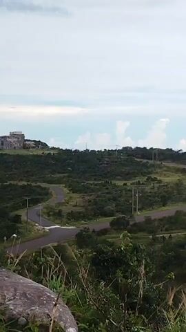 The video opens with a shot of a grassy, hilly landscape with some trees and a road. The camera then pans to the right, revealing a large body of water with land in the distance. The scene then transitions to a wider aerial view of a coastline with more water and land, including what appears to be a man-made structure extending into the water. The video continues to pan and zoom out, showing more of the expansive water and distant landmasses, under a cloudy sky. The overall environment appears to be coastal and natural, with some signs of human development.