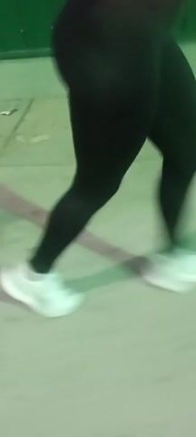 A person with dark leggings and white athletic shoes is walking on concrete. They are also wearing a pink top, partially visible. The video is shaky and focuses primarily on the lower half of the person. The background is a blurry mix of green and grey, likely a wall or building. The camera follows the person’s movement as they walk.