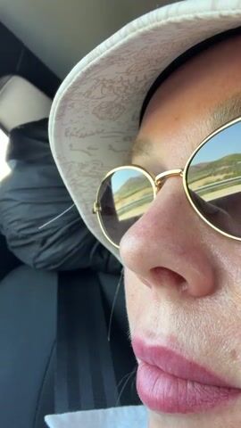 The video shows a close-up, side profile of a person wearing a white hat and gold-rimmed sunglasses, looking out a car window. The reflection in the sunglasses shows a road and a hillside. The person's face is illuminated, and their lips are painted a reddish-pink color. The background is out of focus, but appears to be the interior of a vehicle.